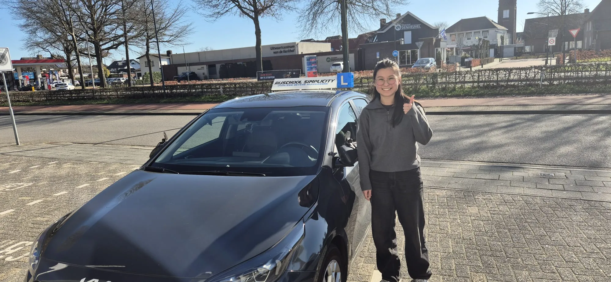 Geslaagde leerling steekt haar duim op naast de lesauto van Rijschool Simplicity